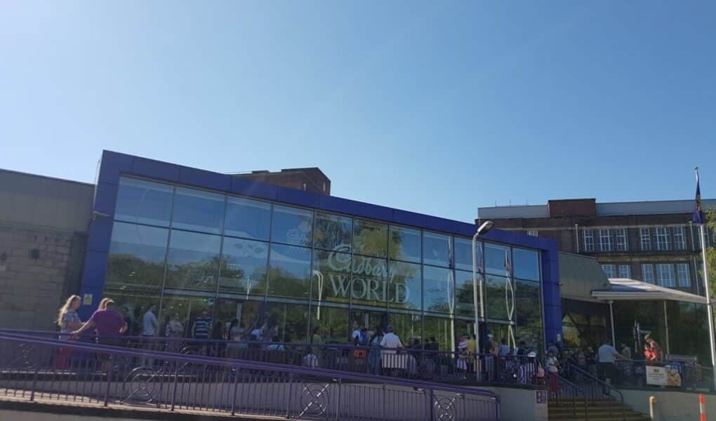 A modern glass-fronted building with “Cadbury World” written on the window. A crowd of people stands outside by a purple railing under a clear blue sky. Some greenery is reflected in the glass. A few lamp posts and nearby buildings are visible.