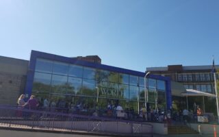 A modern glass-fronted building with “Cadbury World” written on the window. A crowd of people stands outside by a purple railing under a clear blue sky. Some greenery is reflected in the glass. A few lamp posts and nearby buildings are visible.