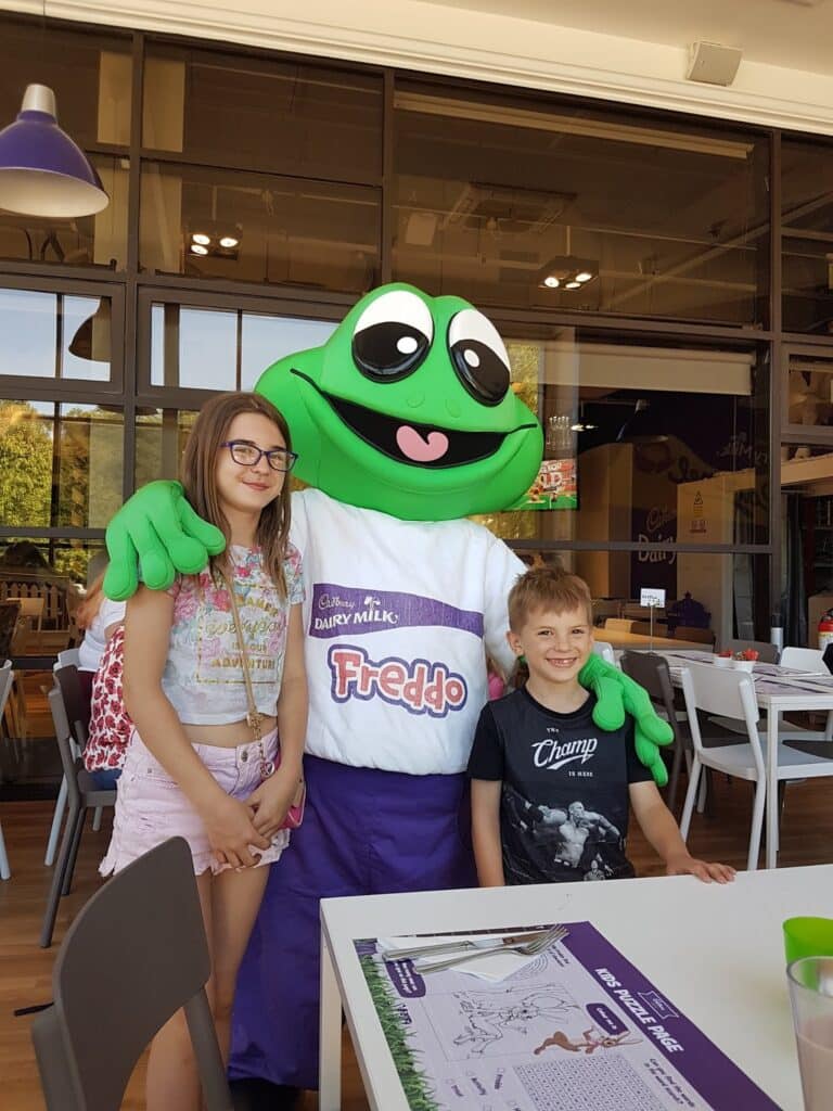 A person in a large green frog costume with big eyes and a "Freddo" shirt stands inside a café, smiling and posing with a young girl in glasses and shorts and a young boy in a T-shirt. Tables and chairs are visible in the bright, modern setting.