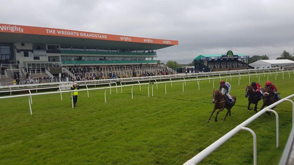 Three jockeys race horses on a green grass track past white railings, nearing a finishing line. Spectators watch from “The Wrights Grandstand” building on the left under a cloudy sky. A race official in yellow is standing on the field near the rail.