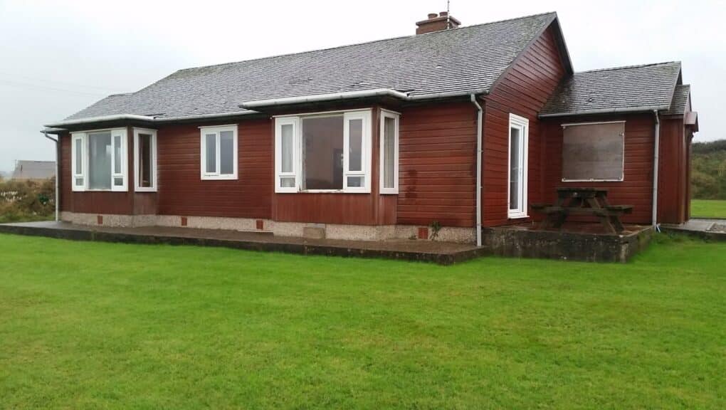 A single-storey brown wooden house with a grey shingled roof, white-framed windows, and a chimney. The house sits on a concrete foundation and is surrounded by a neatly mown green lawn. There is a wooden picnic table on the right side.