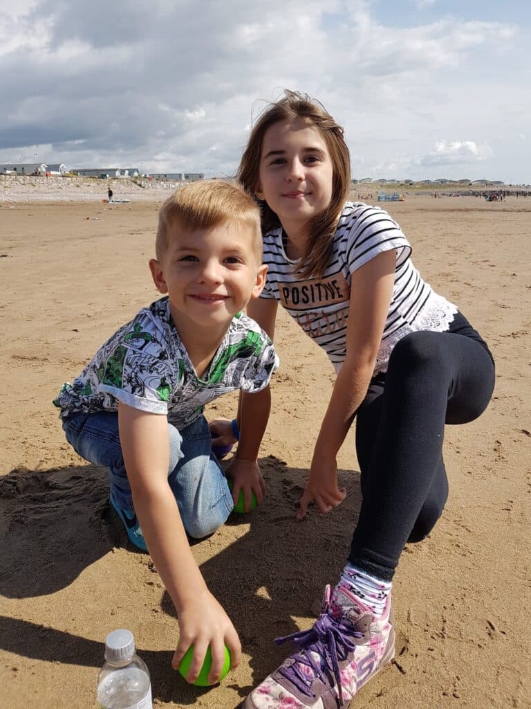 Two children, a boy and a girl, crouch on a sandy beach smiling at the camera. The boy wears a patterned shirt and jeans, the girl wears a striped shirt and black leggings with pink trainers. A water bottle is on the sand. The sky is partly cloudy.