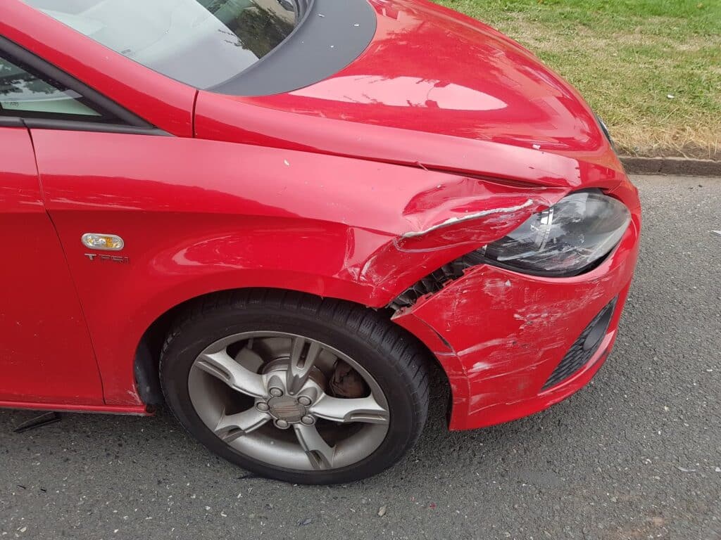 A red car with significant damage to the front left wing and bumper; the headlamp is scratched, and the panel is dented and cracked. The car is parked on a grey road beside grass. The wheel and tyre appear undamaged.