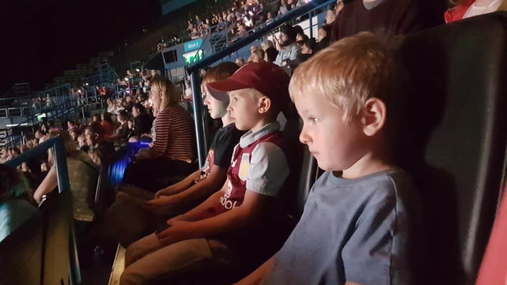Three young children sit in a row of theatre or stadium seats, watching an event. The children look focused and serious. The venue is dimly lit, with other audience members visible in the background and seating stretching up into darkness.