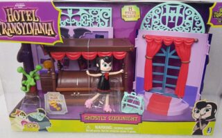 Hotel Transylvania “Ghostly Goodnight” toy set in a box showing a bed, suitcase, plant, balcony window, and a Mavis doll with arms outstretched. The packaging is purple with cartoon graphics and Hotel Transylvania logo at the top left.