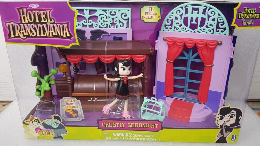 A Hotel Transylvania "Ghostly Goodnight" playset in packaging includes a Mavis doll, a purple bed with red curtains, a blue gate, a green plant, and accessories. The box is purple with Hotel Transylvania branding and animated Mavis artwork.
