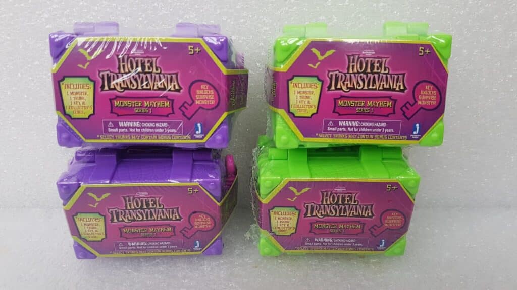Four “Hotel Transylvania Monster Mayhem” surprise toy cases, two green and two purple, are stacked in pairs against a white textured background. Each case features a magenta label with yellow text, highlighting they are suitable for ages 5+.