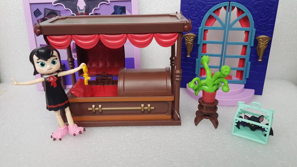 A toy vampire girl with pink monster slippers holds a golden key next to a brown and red toy coffin. Behind her are purple and blue toy doors, a green potted plant on a stand, and a black bat in a turquoise pet carrier.