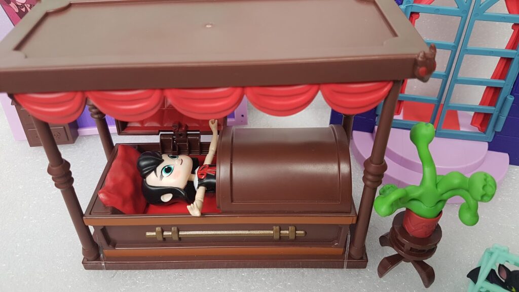A gothic-style toy doll lies in a brown canopy bed with red curtains and bedding in a dolls' house setting. The doll has pale skin, black hair, and is partially covered by a curved lid. Nearby is a green potted plant and colourful wall décor.