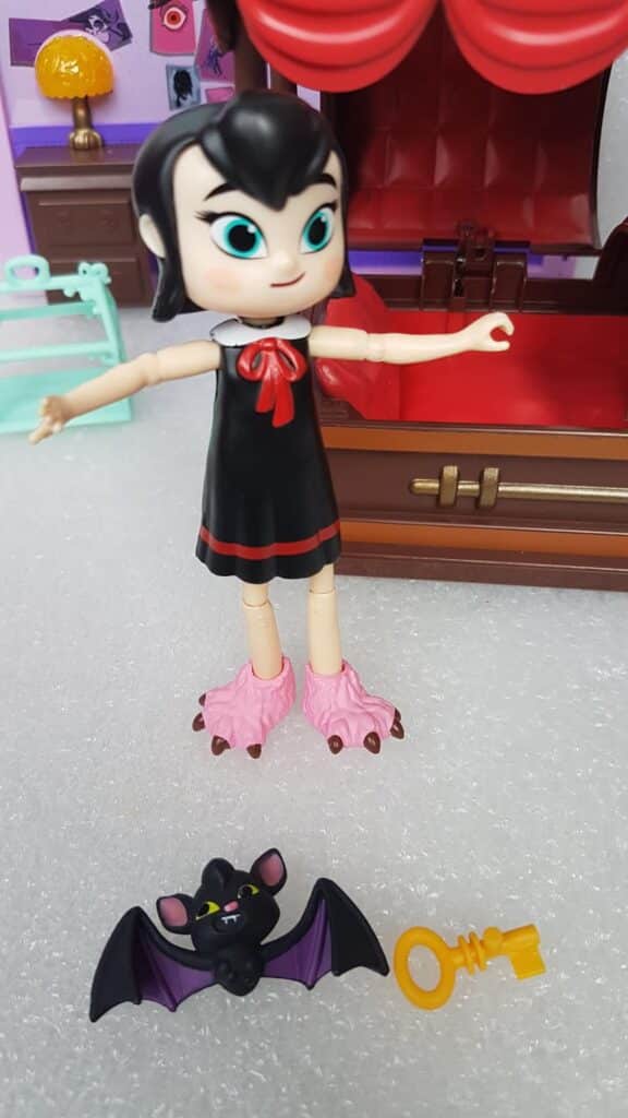 A doll with pale skin, black hair, and blue eyes wears a black dress with a red bow and pink monster slippers. In front of her are a small bat toy with purple wings and a yellow key. Behind her is a brown coffin-shaped bed and purple bedroom décor.