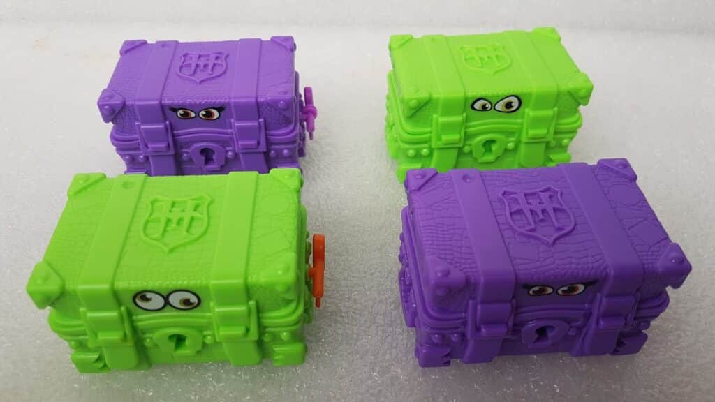 Four toy treasure chests with cartoon eyes, two mostly green with purple trim, and two mostly purple with green trim, sit on a white textured surface. Each chest has a moulded crest, keyhole, and detailed texture, giving a playful and quirky appearance.