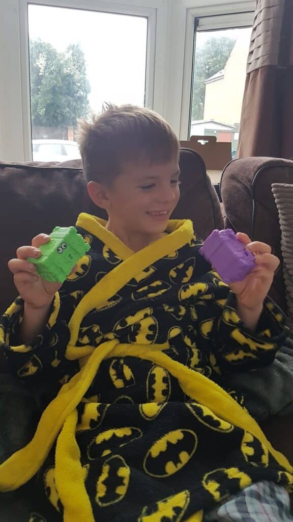 A young boy in a black and yellow Batman dressing gown sits on a sofa, smiling while holding a green toy in one hand and a purple toy in the other. The room is softly lit by daylight from a large window behind him.