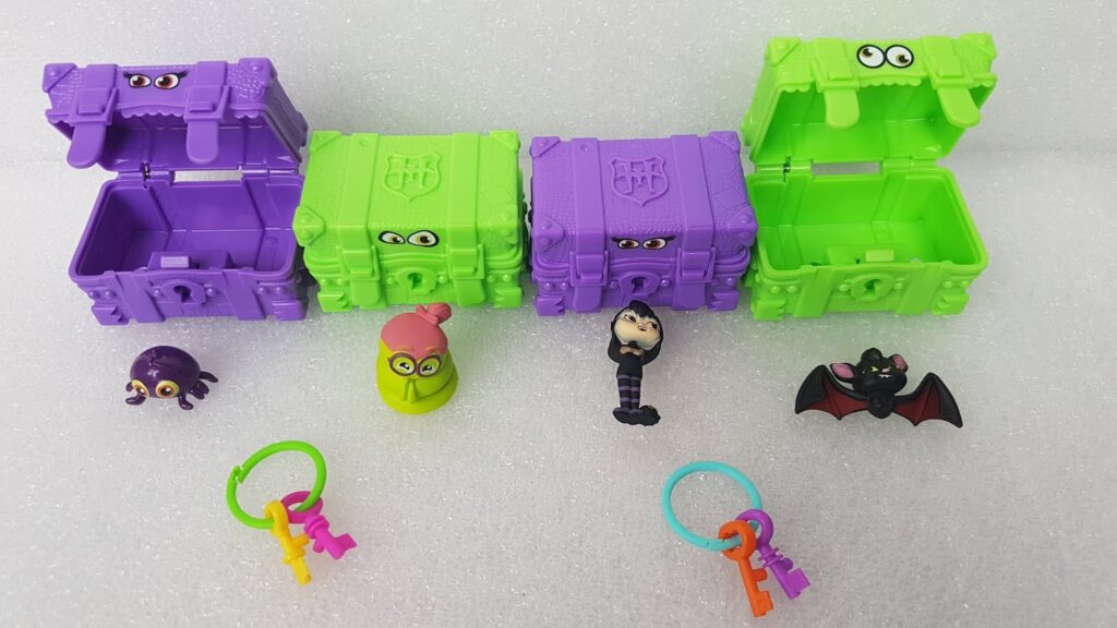 Three colourful toy treasure chests (two purple, one green) with monster eyes, stand open on white foam. In front are four small monster figurines—a spider, a green creature, a worm, and a bat—plus two keyrings with pink, purple, and orange keys.