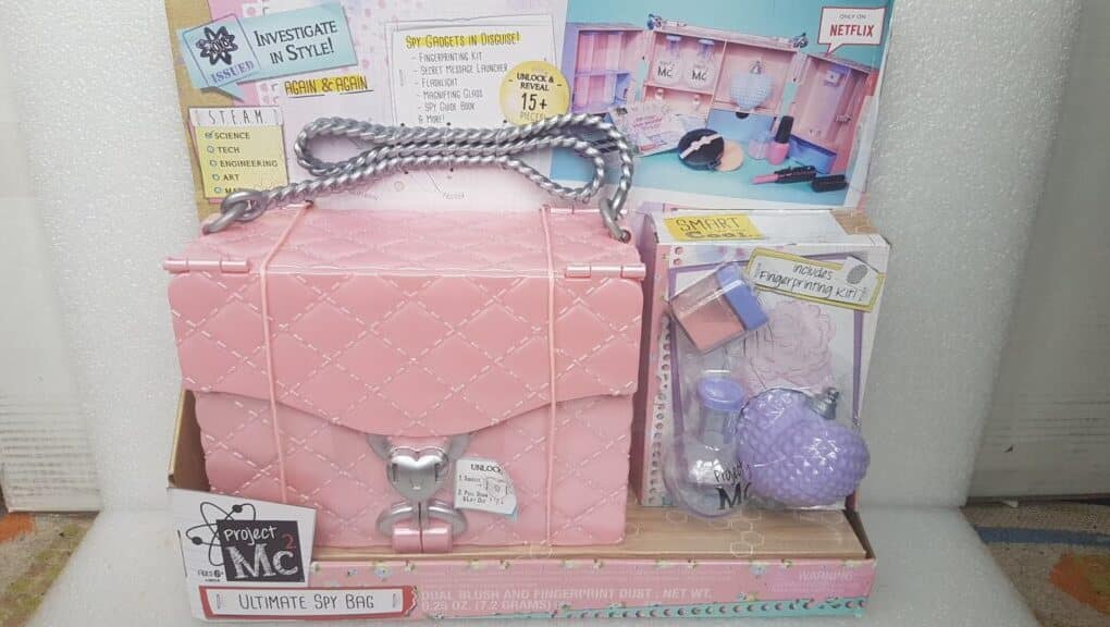 A Project Mc² Ultimate Spy Bag toy set is displayed in its packaging. The set includes a pink quilted bag with a silver lock and handle, and spy-themed tools. The packaging is colourful, showing accessories and instructions for children aged 6 and up.