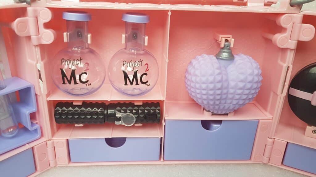A pink plastic toy science kit with two clear round bottles labelled "Project Mc²," a purple textured heart-shaped container, black cylindrical pieces, and two blue drawers. The compartments are neatly arranged inside the opened kit.