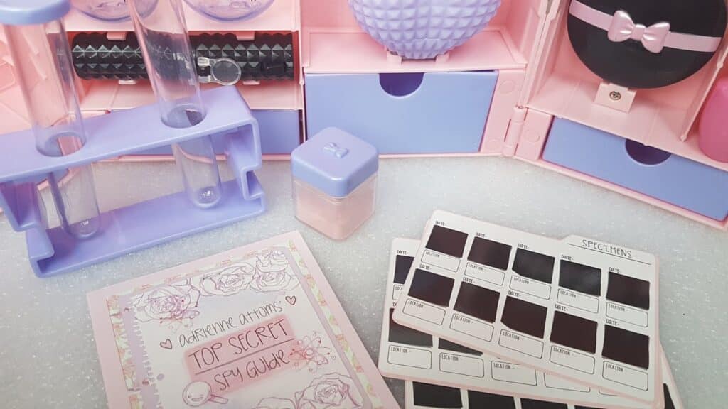 A pastel-themed spy kit with a lavender test tube rack, two test tubes, a pink and purple case, a small purple container, a “Top Secret Spy Guide” booklet decorated with flowers, and two sheets labelled “SPECIMENS” holding dark colour samples.