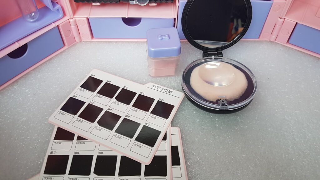 A pastel make-up organiser with drawers is open. In front, there are two shade sample cards, a small purple powder container, and a round compact with a mirror and sponge, all arranged on a light, textured surface.