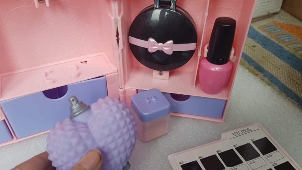 A hand holds a purple, textured spray bottle in front of a pink and purple toy make-up set. The set includes small drawers, a black compact with a pink bow, a pink bottle, a purple container, and a make-up palette labelled "SPLATMAT.