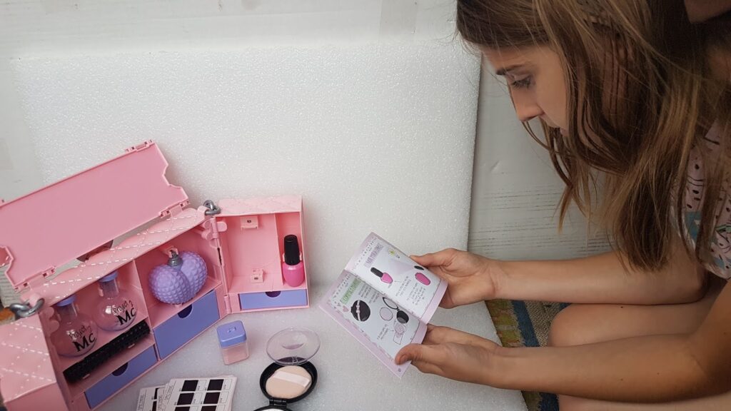 A young girl sits on the floor, closely examining an instruction booklet while a pink toy make-up set with various miniature beauty items, including bottles and palettes, is open beside her against a white background.