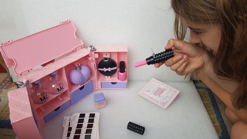 A young girl plays with a pink toy make-up set that includes pretend perfume bottles, a black and pink lipstick, nail varnish, and an eyeshadow palette. She holds up a large toy lipstick, examining it closely, with the open make-up case in front of her.