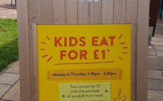 A wooden sandwich board outside, displaying a sign for "Cookhouse and Pub." The yellow poster reads "Kids eat for £1," valid Monday to Thursday, 3.30–5.30pm, with the purchase of an adult main course. Simple food illustrations surround the text.