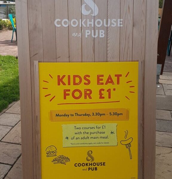A wooden sandwich board outside, displaying a sign for "Cookhouse and Pub." The yellow poster reads "Kids eat for £1," valid Monday to Thursday, 3.30–5.30pm, with the purchase of an adult main course. Simple food illustrations surround the text.