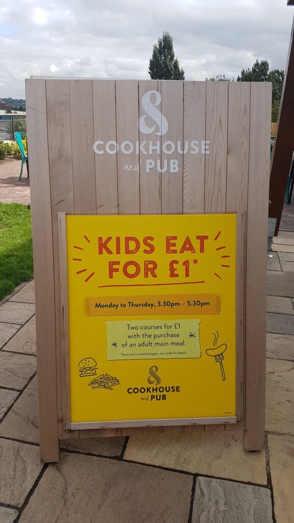 A wooden sign for Cookhouse and Pub displays a yellow poster reading, "KIDS EAT FOR £1*," valid Monday to Thursday, 3:30 pm–5:30 pm, with two courses for £1 with the purchase of an adult main course. Simple food icons decorate the poster.