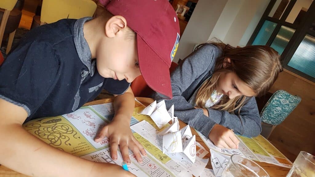 A boy in a red cap and a girl with long hair sit at a table, focused on drawing or writing on colourful activity sheets. Several paper fortune tellers are on the table between them. They appear engaged in a creative or educational activity indoors.