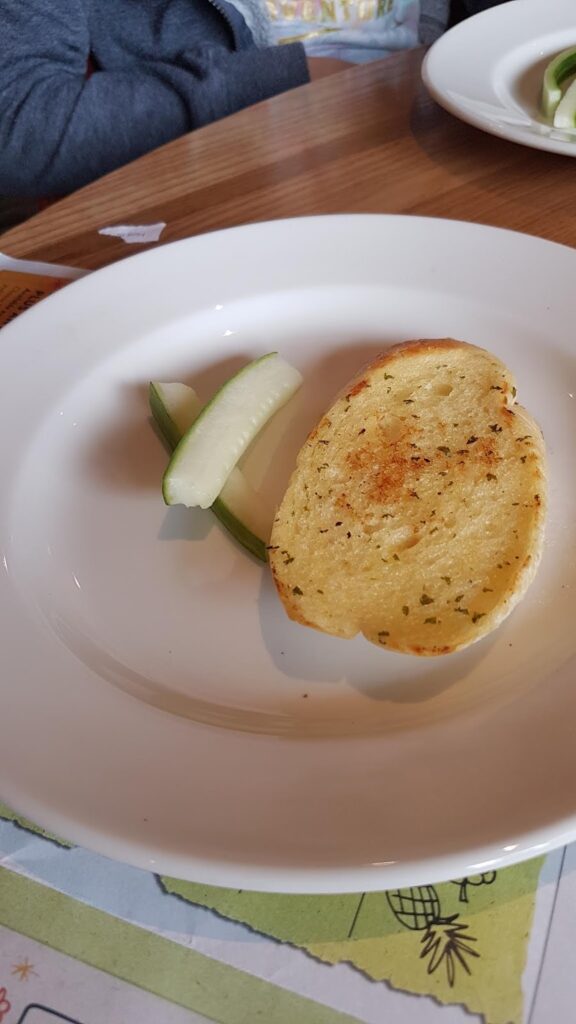 A white plate holds a slice of toasted garlic bread with herbs and two spears of cucumber. The plate is on a wooden table, and part of another plate and placemat are visible in the background.