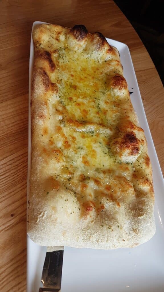 A rectangular flatbread with a golden, bubbly, and slightly charred crust, topped with melted cheese and herbs, sits on a white rectangular plate atop a wooden table. A metal utensil is partially visible under the plate.
