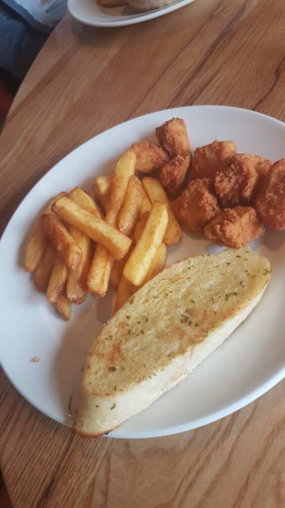 A white plate on a wooden table holds golden chips, crispy breaded chicken pieces, and a slice of garlic bread with herbs. The food is arranged separately, creating a balanced, casual meal.