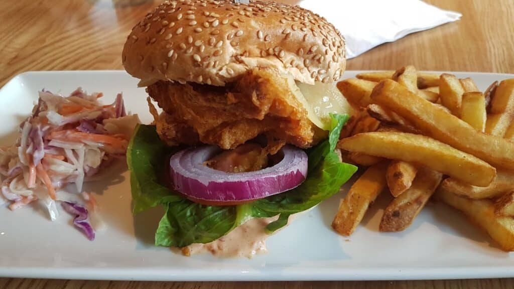 A rectangular white plate holds a battered fish sandwich with lettuce, red onion, sauce, and a sesame seed bun, accompanied by a serving of golden chips and a portion of creamy coleslaw. The background shows a wooden table.