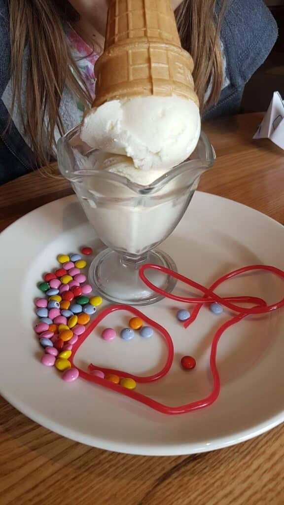 A glass bowl with vanilla ice cream topped by an upside-down cone sits on a white plate. On the plate are colourful round sweets and two long red jelly sweets. A person with long brown hair and a grey shirt is partially visible in the background.