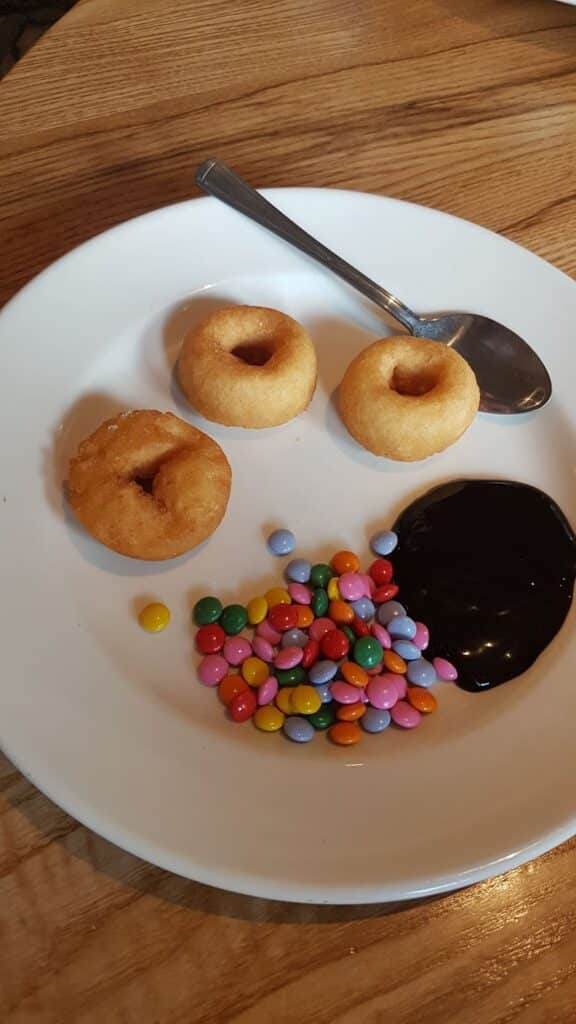 A white plate on a wooden table holds three mini doughnuts, a pile of colourful candy-coated chocolates, a spoon, and a swirl of chocolate sauce. The arrangement resembles a smiling face.