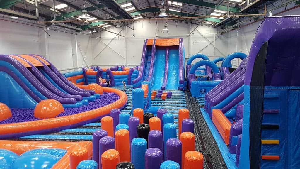 A large indoor inflatable assault course features bright blue, orange, and purple structures, including slides, climbing walls, and pillars, all under a high, industrial-style ceiling with exposed beams and lights. A few people can be seen playing inside.