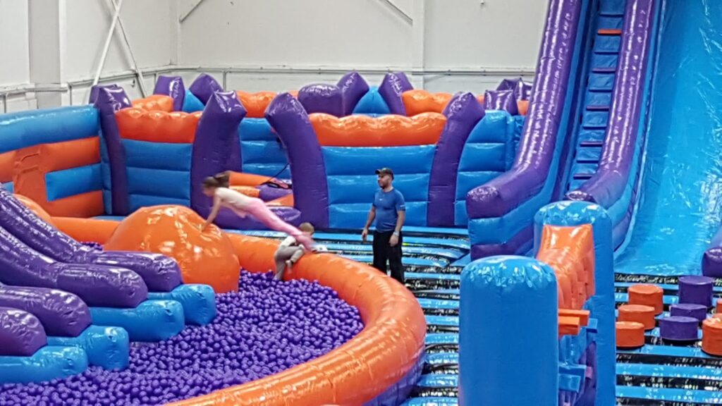 A young girl in pink leaps onto an orange inflatable obstacle in an indoor play area filled with purple plastic balls. A man in a blue shirt watches nearby. The play area features large purple, orange, and blue inflatable structures and slides.