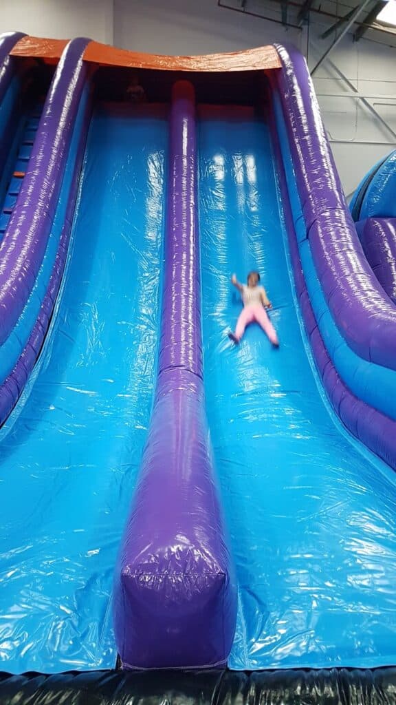 A child in pink clothing slides down a large inflatable slide with blue and purple lanes inside an indoor play centre. The child is slightly blurry, indicating motion. The slide has barriers on each side and a partition in the middle.