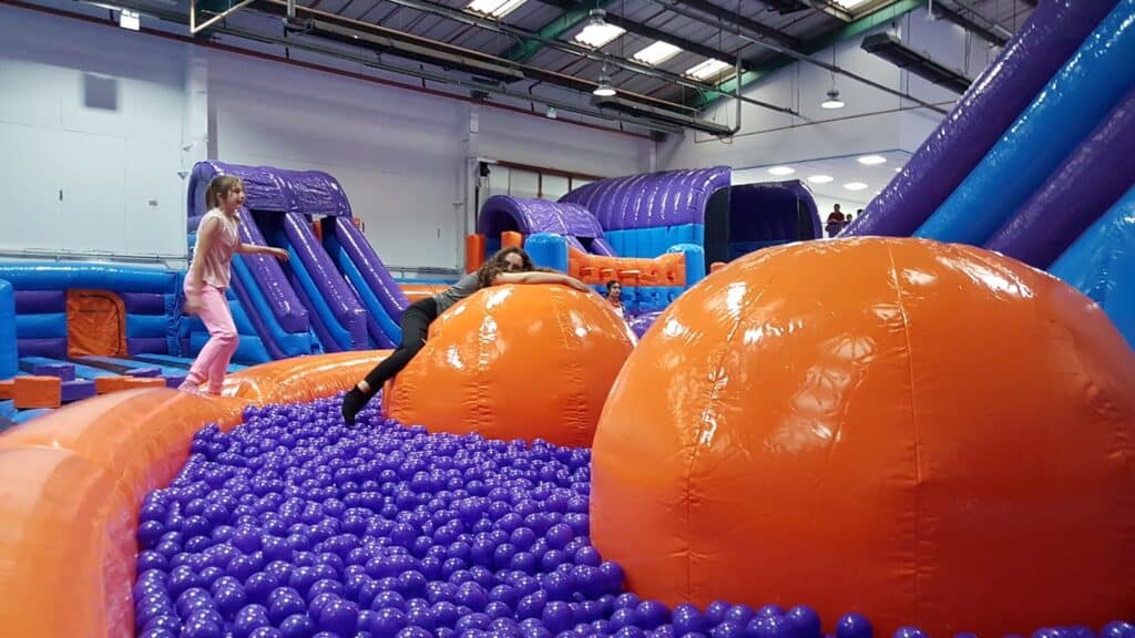 Two children play on a large indoor inflatable assault course with bright orange spheres and a pit of blue plastic balls. The area is surrounded by purple and blue inflatables, with slides and arches visible under a high ceiling with skylights.