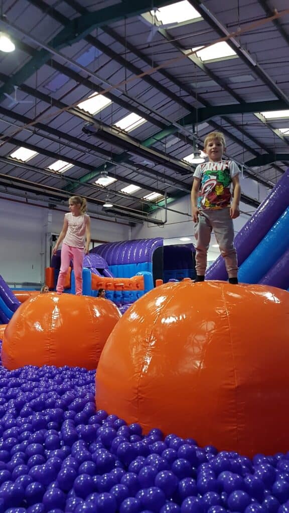 Two children stand on large, shiny orange inflatable spheres surrounded by a sea of small purple balls inside an indoor play centre. The background shows blue and purple inflatable structures and a high ceiling with exposed beams and lights.