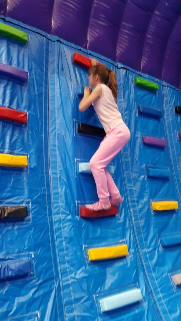 A young girl in a pink outfit climbs a blue inflatable wall with colourful horizontal blocks for steps. The indoor wall has a purple, arched top, and she appears focused as she reaches for a red step near the upper section.