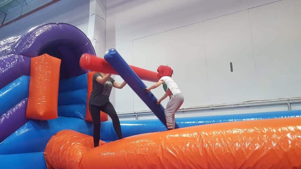Two people wearing helmets stand on an inflatable orange platform, each holding a long foam bat and playfully jousting. The scene is indoors, with colourful inflatable structures in blue, orange, and purple in the background.