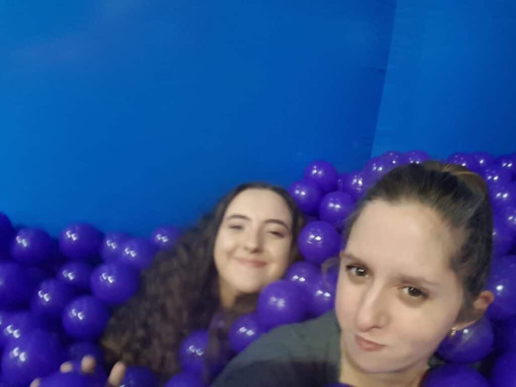 Two young women are in a pool filled with purple plastic balls, set against a blue background. One woman in front has brown hair tied back, with a neutral expression. The other, behind her, has curly hair and is smiling slightly.