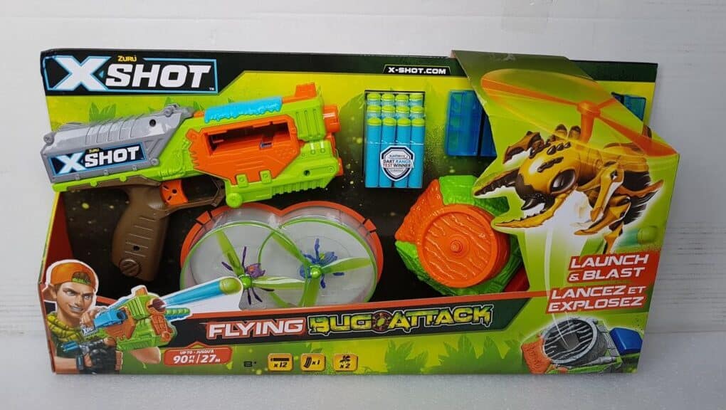 A toy set in a colourful box labelled "X-Shot Flying Bug Attack." The set includes a green and orange toy blaster, foam darts, spinning flying bug targets, and a bug-shaped launcher with a yellow insect graphic. The packaging shows a boy aiming the blaster.