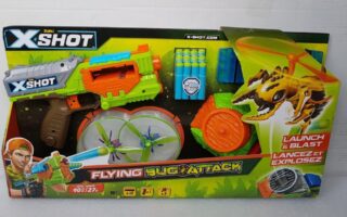 A toy set in a colourful box labelled "X-Shot Flying Bug Attack." The set includes a green and orange toy blaster, foam darts, spinning flying bug targets, and a bug-shaped launcher with a yellow insect graphic. The packaging shows a boy aiming the blaster.