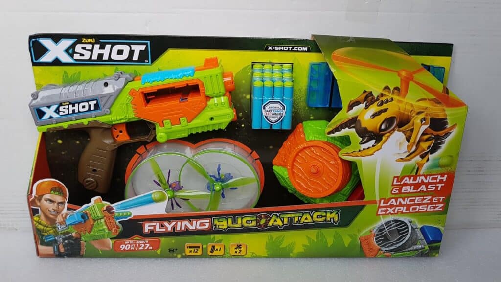 A toy dart gun set called “X-Shot Flying Bug Attack” by Zuru. The packaging shows a green and orange dart blaster, foam darts, a launcher, spinning bug targets, and action graphics, with text reading “Launch & Blast” in English and French.