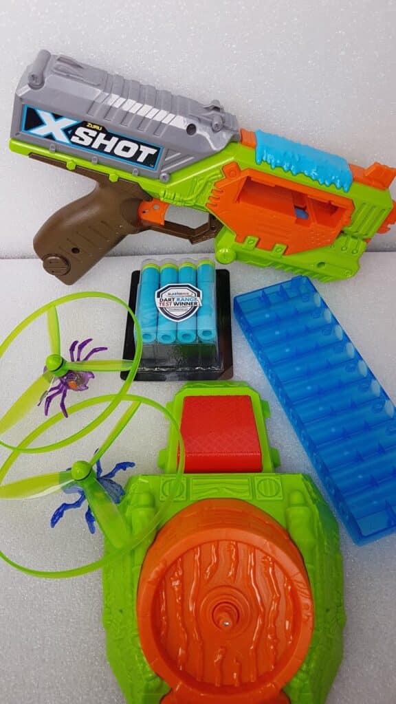 Colourful toy set on a white surface: a green and orange toy blaster labelled "X-Shot," blue foam darts in a holder, a blue plastic piece with slots, a green and orange spinning disc launcher, and two plastic discs, one with purple propellers.