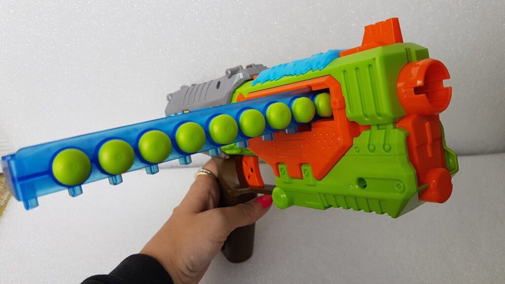 A hand holding a colourful toy gun with a green, orange, blue, and brown body. The gun has a blue chamber loaded with multiple green foam balls, ready to be fired. The background is a plain, light-coloured surface.