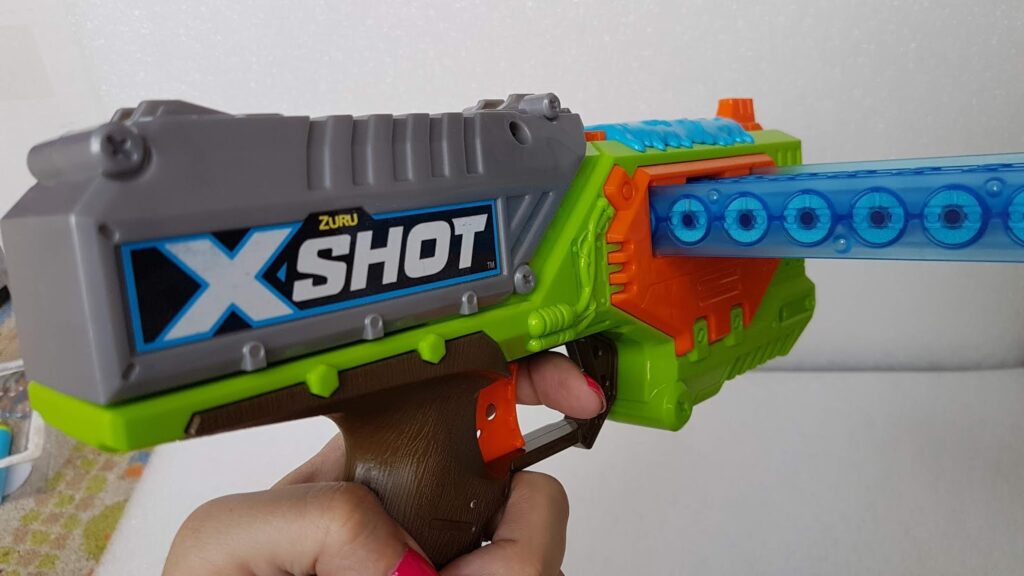 A hand holds a toy blaster labelled “X-Shot” in bold letters. The blaster is grey, green, orange, and brown, with a blue transparent rotating dart chamber loaded with foam darts. The background is plain white.