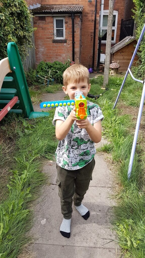 A young boy stands on a garden path pointing a colourful toy blaster at the camera. He is wearing a dinosaur-print shirt, green trousers, and socks. Behind him is a green slide, a swing set, and a red-brick house with grass and plants around.