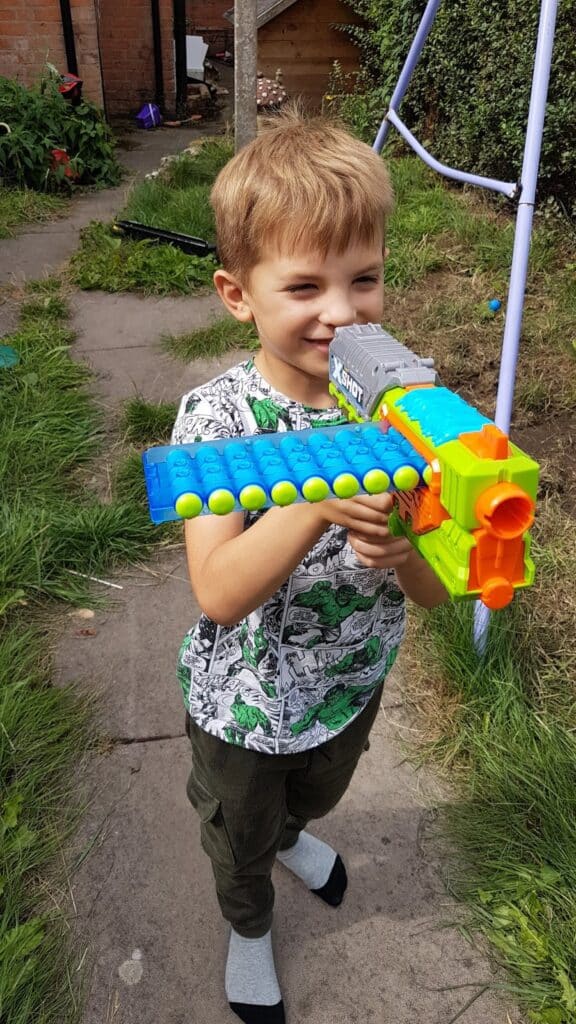A young boy with blond hair stands on a garden path, smiling and holding a colourful toy blaster loaded with foam balls. He wears a white T-shirt with green patterns, green trousers, and mismatched socks. Grass and garden equipment are visible in the background.